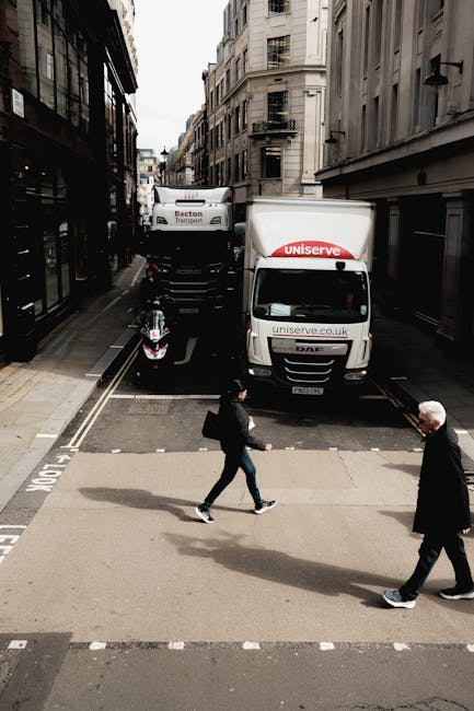 An urban street scene showing a narrow road bordered by tall, multi-storey buildings with classic architectural features, including balconies and large windows. In the foreground, two pedestrians are crossing a zebra crossing; one is a young woman carrying a backpack and wearing casual clothing, while the other is an older man in a dark coat. Behind them, a white delivery van with the logo of Uniserve and the URL uniserve.co.uk is parked partly on the pavement, close to a sidewalk curbed by metal barriers. Further back, a larger truck with visible side markings is parked opposite the van, and a black scooter is parked near the curb adjacent to the vehicles. The scene likely depicts a typical residential or commercial area in Fulham, with street parking and operational loading zones, relevant to house removals or home relocation services. Man With a Van Fulham occasionally handles such logistics, including vehicle loading and unloading processes for moving and furniture transport.