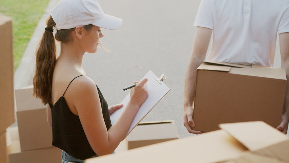 A woman with long brown hair tied in a ponytail, wearing a white cap, black sleeveless top, and holding a clipboard with a pen, is standing outside a property during a house move. She appears to be taking notes or inventory as part of the packing and moving process. In front of her, a man wearing a white T-shirt is holding a medium-sized cardboard moving box, with several other boxes stacked nearby. The scene is set on a paved driveway or sidewalk, with the background showing green grass and natural outdoor lighting. Inside or near the property, additional cardboard boxes are visible, some covered with plastic film or packing materials. The presence of moving equipment such as trolleys, blankets, or straps is not clearly visible, but the individuals are engaged in organizing or preparing items for transport, reflecting a home relocation or furniture transport activity. This scene aligns with professional removals services offered by Man With a Van Fulham, capturing the typical staging of a house removal.