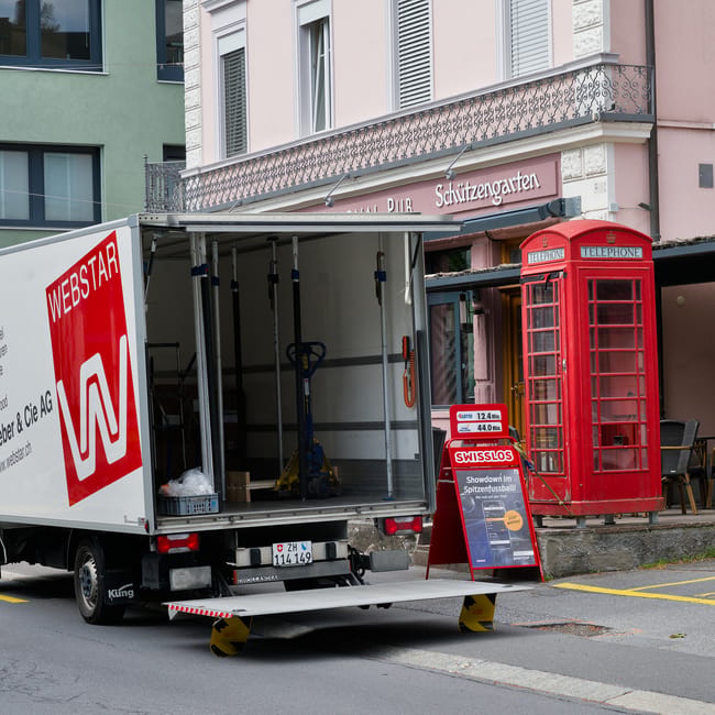An urban street scene showing a narrow road bordered by tall, multi-storey buildings with classic architectural features, including balconies and large windows. In the foreground, two pedestrians are crossing a zebra crossing; one is a young woman carrying a backpack and wearing casual clothing, while the other is an older man in a dark coat. Behind them, a white delivery van with the logo of Uniserve and the URL uniserve.co.uk is parked partly on the pavement, close to a sidewalk curbed by metal barriers. Further back, a larger truck with visible side markings is parked opposite the van, and a black scooter is parked near the curb adjacent to the vehicles. The scene likely depicts a typical residential or commercial area in Fulham, with street parking and operational loading zones, relevant to house removals or home relocation services. Man With a Van Fulham occasionally handles such logistics, including vehicle loading and unloading processes for moving and furniture transport.