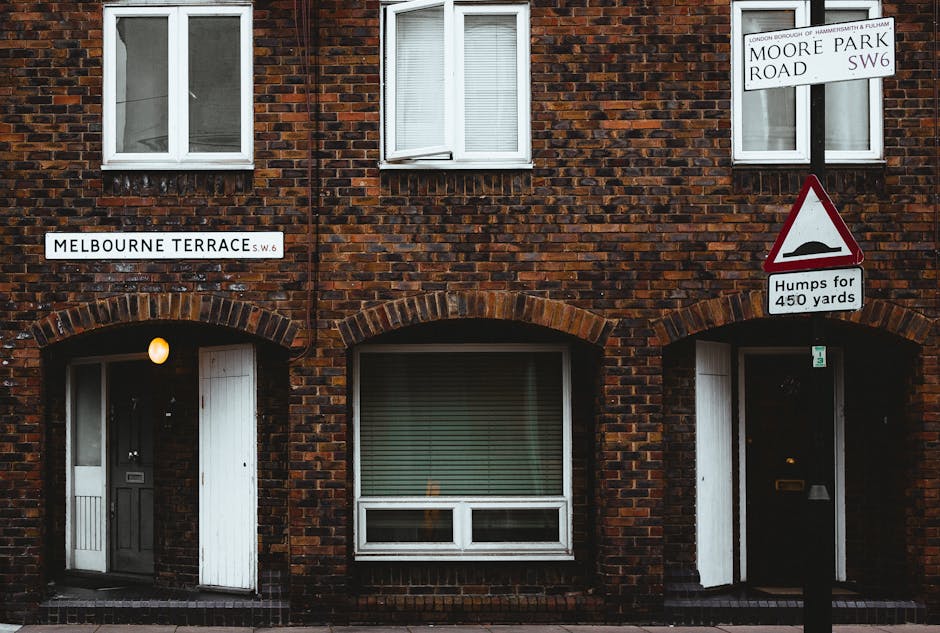 A brick residential building on Melbourne Terrace with a double-story structure displaying three white-framed windows, two on the upper floor and one on the ground floor. The ground floor features two arched doorways with dark-colored entrances, one of which is partially open, revealing a light fixture inside, while the other is closed. A large window with horizontal blinds is situated between the doorways. On the street corner, there are two road signs: one indicating the street name as Moore Park Road SW6 and another warning of upcoming road humps for 450 yards. The scene is captured during daylight with natural lighting, suitable for documenting house removals or moving activities, as part of a professional property relocation service like Man With a Van Fulham. The street scene provides context for the logistics involved in local house removals, including access points and planning considerations for furniture transport and packing during a home relocation. 