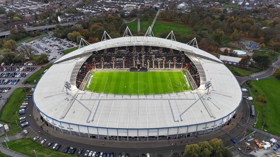 The image shows an empty football stadium with a bright green grass pitch, surrounded by multiple tiers of red and pink seats. Above the seating areas, a large digital scoreboard hangs from the stadium's roof, which features a white steel framework supporting a clock. The sky is clear with natural daylight illuminating the scene. In the background, tall modern buildings are visible beyond the stadium's upper level, indicating an urban location. The stadium appears to be prepared for a match day, with no people present on the field or in the stands. This setting could be relevant in the context of a house removal or relocation project, such as preparing for stadium-based events or managing transport logistics around large venues, an aspect relevant to home relocations in Fulham related to match days, coordinated by Man With a Van Fulham, a professional removals service specializing in house moves and transportation.