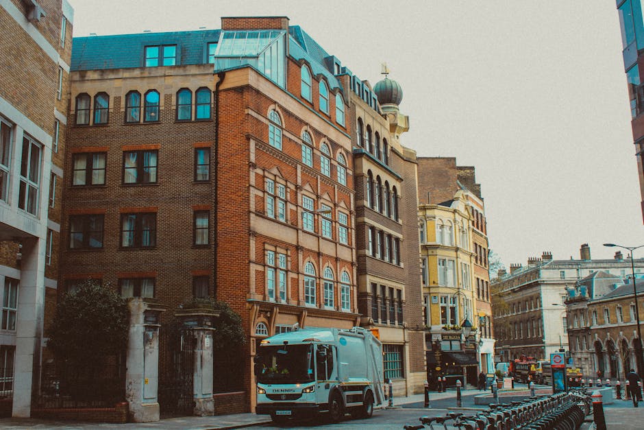 Image depicting a city street scene with a white man with a van Fulham vehicle parked at the curb in front of a row of historic multi-storey buildings with brick facades and large windows. The van is