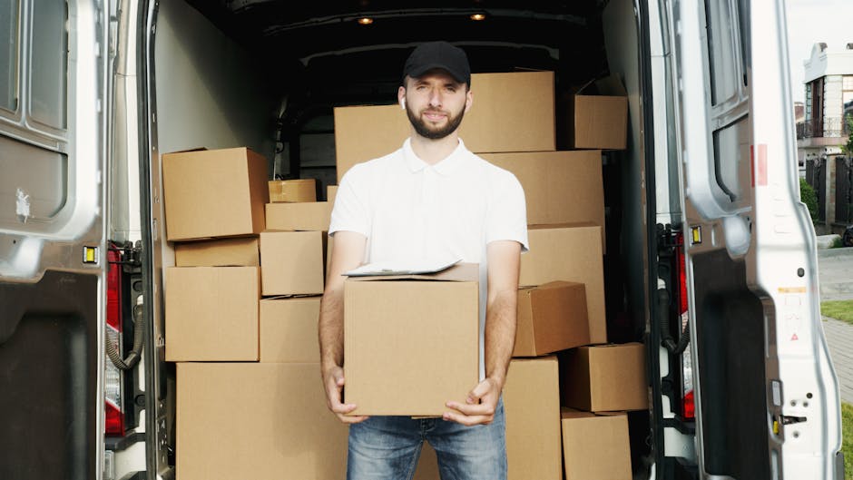 A man with a beard dressed in a white polo shirt, dark jeans, and a black cap stands inside the open rear of a white van, which is loaded with numerous packed cardboard boxes of various sizes. The boxes are stacked neatly against the van's interior walls, some wrapped in plastic or packing materials, indicating careful packing for a house move. The man is holding a medium-sized cardboard box with both hands, positioned at waist level, and has a neutral expression as he looks at the camera. The background shows a residential street with pavement and houses, suggesting that the loading process is taking place at a home address in Fulham. The image captures a moment of furniture transport and packing during the home relocation process, illustrating the services provided by Man With a Van Fulham in a professional and straightforward manner, suitable for house removals and moving logistics.