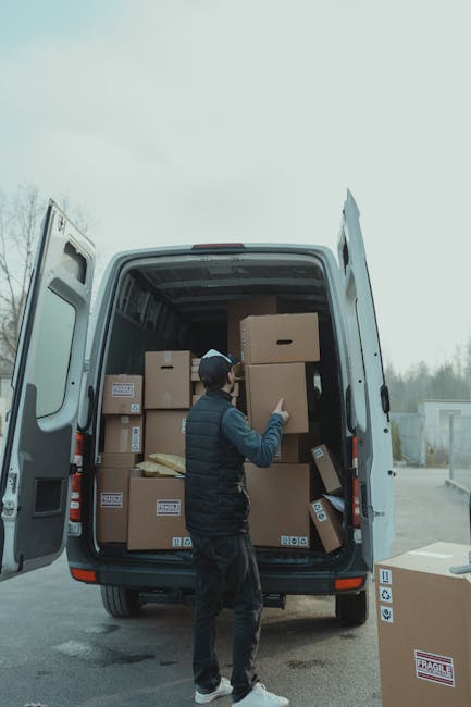 A man with a beard dressed in a white polo shirt, dark jeans, and a black cap stands inside the open rear of a white van, which is loaded with numerous packed cardboard boxes of various sizes. The boxes are stacked neatly against the van's interior walls, some wrapped in plastic or packing materials, indicating careful packing for a house move. The man is holding a medium-sized cardboard box with both hands, positioned at waist level, and has a neutral expression as he looks at the camera. The background shows a residential street with pavement and houses, suggesting that the loading process is taking place at a home address in Fulham. The image captures a moment of furniture transport and packing during the home relocation process, illustrating the services provided by Man With a Van Fulham in a professional and straightforward manner, suitable for house removals and moving logistics.