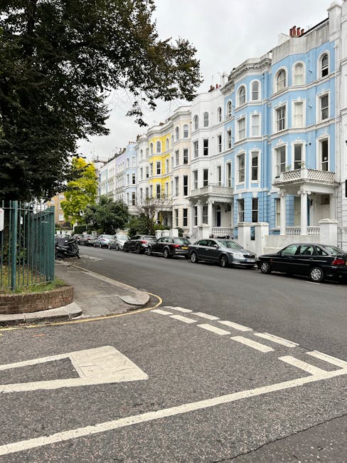 Image of a residential street in Sands End with a row of multi-story Victorian-style buildings painted in pastel colors including white, yellow, and light blue. Parked cars line the curb beside a smooth asphalt road, which features white dashed parking lines and a pedestrian crossing with striped markings. On the left side, a green metal fence borders a sidewalk with a small tree and some shrubs visible nearby. The sky is overcast, casting diffuse light over the scene. In the context of house removals and moving logistics, this street setting suggests a typical urban environment where furniture transport and packing activities might take place, with ample on-street parking and accessible building entrances, suitable for home relocation processes handled by services such as Man With a Van Fulham.