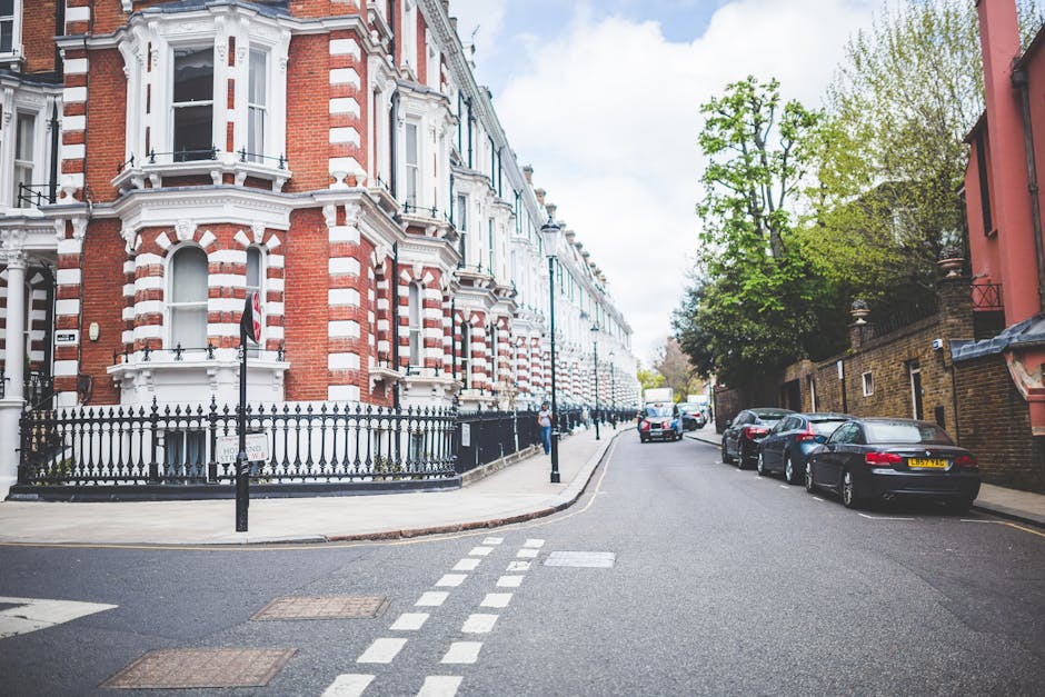 A street scene in Sands End near Fulham Palace featuring a white residential building with multiple levels, ornate window frames, and a small balcony on the second floor. In front of the building, there is a narrow sidewalk with pedestrians walking, some carrying small bags. Parked along the curb are a black van and a dark-colored car, both positioned near an area where loading and unloading might occur. A mid-sized tree with green foliage partially obscures part of the building, casting shadows on the pavement. The street is marked with dashed lines indicating parking spaces, and the sky above is mostly clear with a few scattered clouds. The environment suggests a quiet residential area suitable for house removals and furniture transport, with some visible signage and street lamps indicating an accessible urban neighbourhood. Occasionally, [COMPANY_NAME] may carry out home relocation services in contexts similar to this, supporting efficient moving logistics.